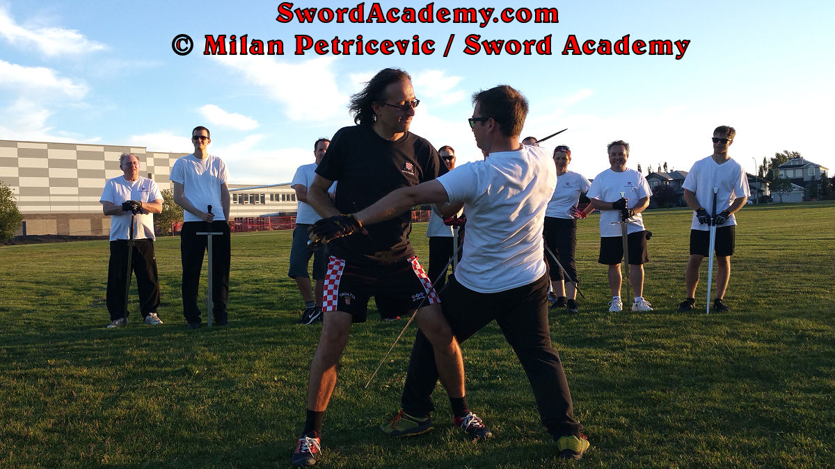 Milan demonstrates during an outdoor class in front of Sword Academy students armored sword exercise / drill using a throw aided by a weapon against the opponent as inspired by historical sources from the German medieval (and renaissance) tradition, part of Sword Academy HEMA / WMA / Martial Arts curriculum.