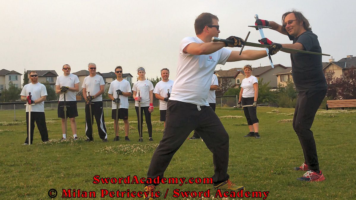 Milan demonstrates during an outdoor class in front of Sword Academy students armored sword exercise / drill using thrust to partially disarm attacking opponent as inspired by historical sources from the German medieval (and renaissance) tradition, part of Sword Academy HEMA / WMA / Martial Arts curriculum.