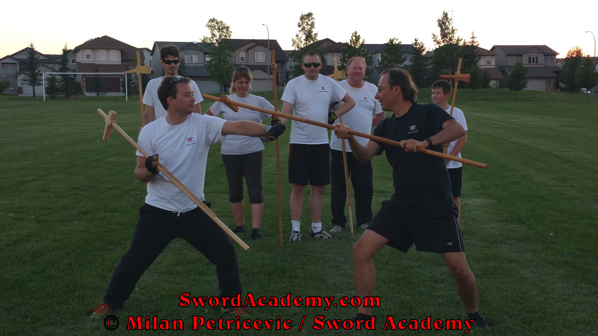 Milan demonstrates during an outdoor class in front of Sword Academy students poleaxe exercise / drill using queue to remove left hand from the shaft and following with the rising strike inspired by historical sources from the French (and German) medieval (and renaissance) tradition, part of Sword Academy HEMA / WMA / Martial Arts curriculum.