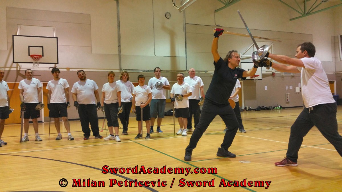 Milan demonstrates during an indoor class in front of Sword Academy students sword and buckler exercise / drill using followup descending thrust to the head from the bind inspired by historical sources from the German medieval and renaissance tradition, part of Sword Academy HEMA / WMA / Martial Arts curriculum.