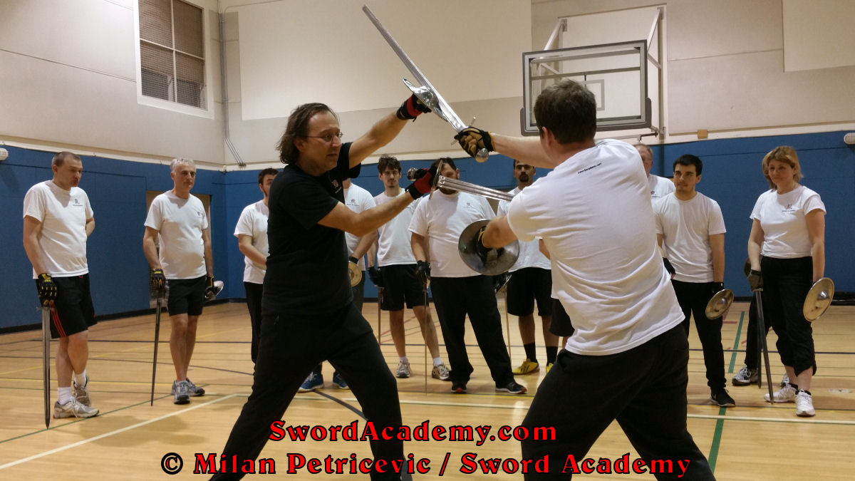 Milan demonstrates during an indoor class in front of Sword Academy students sword and buckler exercise / drill using a separation of sword and buckler to deliver a thrust inspired by historical sources from the German medieval and renaissance tradition, part of Sword Academy HEMA / WMA / Martial Arts curriculum.